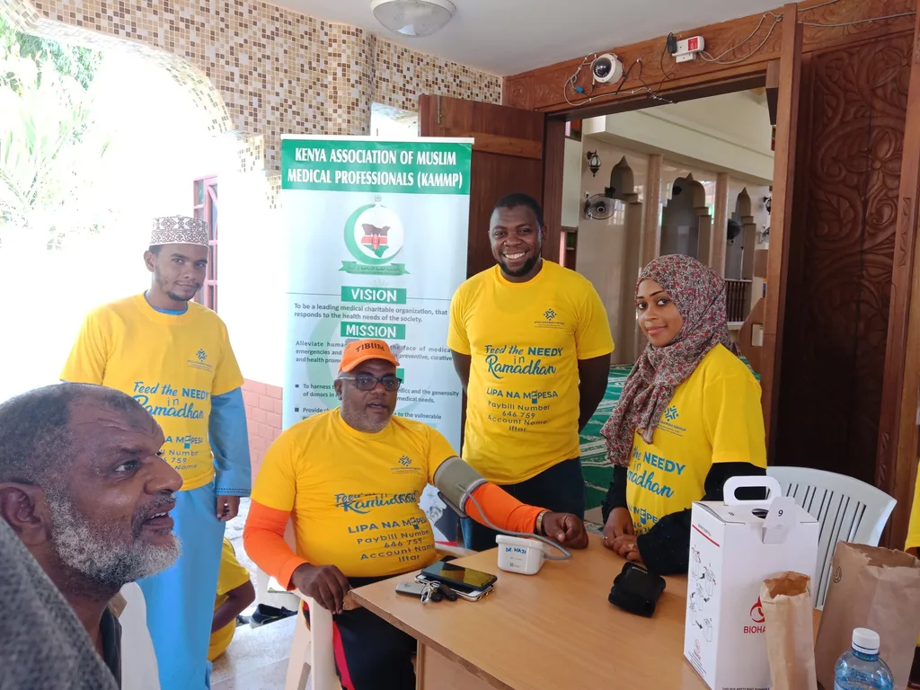 KAMMP volunteers in yellow Ramadan shirts at their community health table beside the association banner, 2019