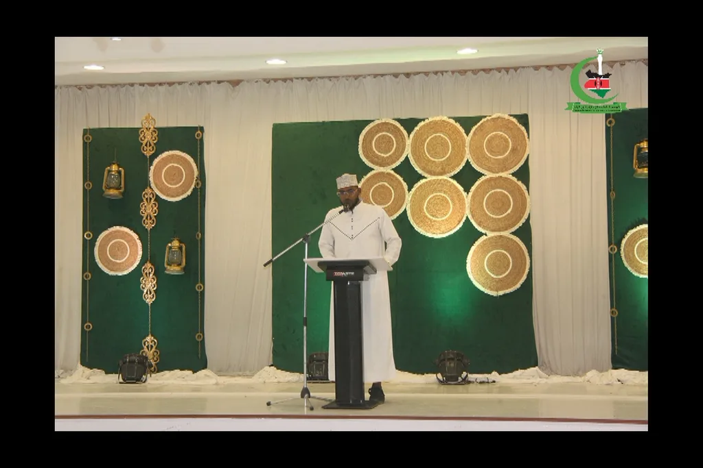 A speaker at the lectern of the Coast Iftar CME, the stage dressed in green and gold, 2024